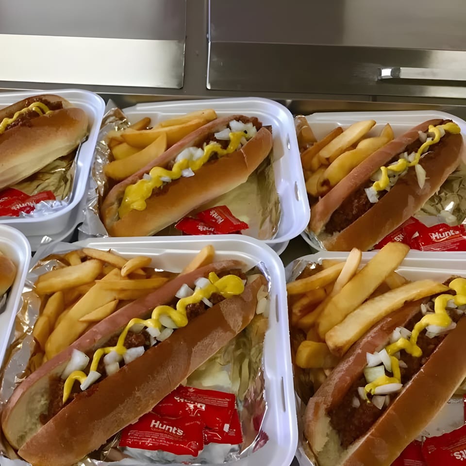 Multiple chili dogs with mustard and onions, served with french fries and ketchup packets in takeout containers.