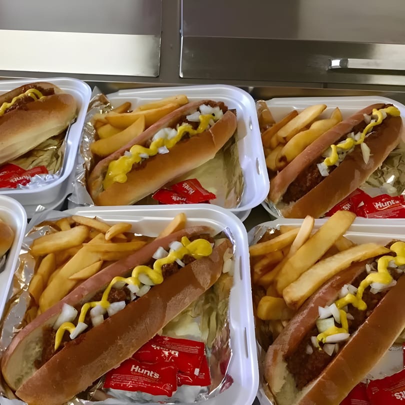 Multiple chili dogs with mustard and onions, served with french fries and ketchup packets in takeout containers.