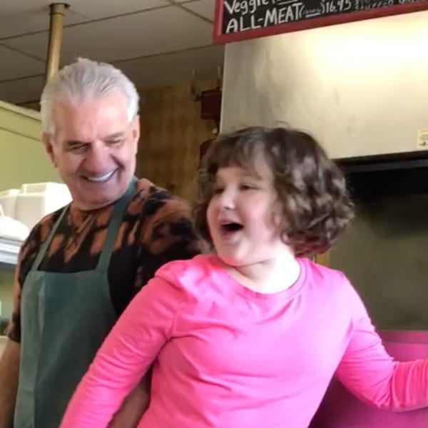 A smiling man in an apron stands behind a young girl in a pink shirt, both laughing.