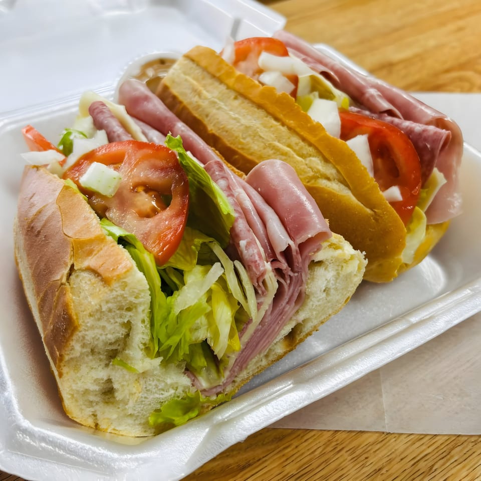 Two Italian subs with deli meat, lettuce, tomatoes, and onions in a white takeout container.