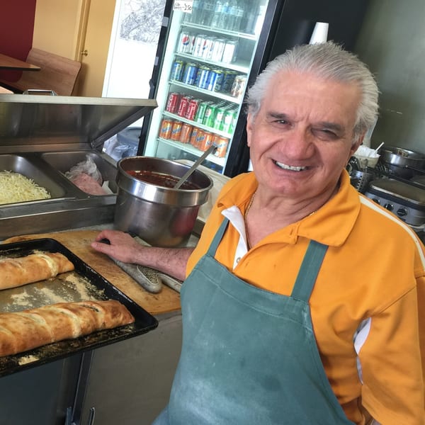 Smiling Vito's Bistro owner in an orange shirt and apron, with calzones and ingredients in the kitchen.